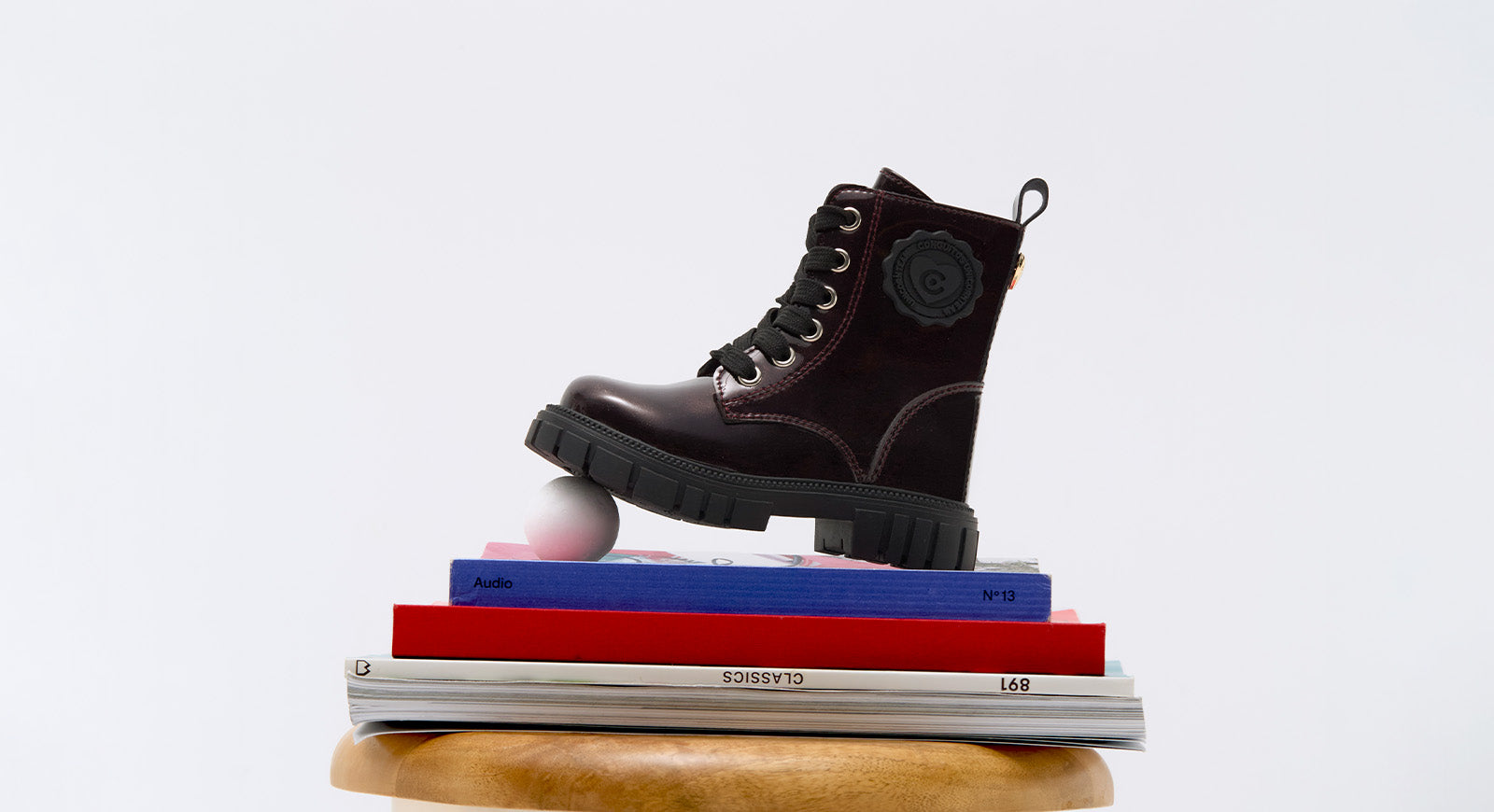 Bota infantil negra con acabado brillante, suela gruesa y cordones, colocada sobre una pila de libros y una pelota blanca, con fondo blanco.