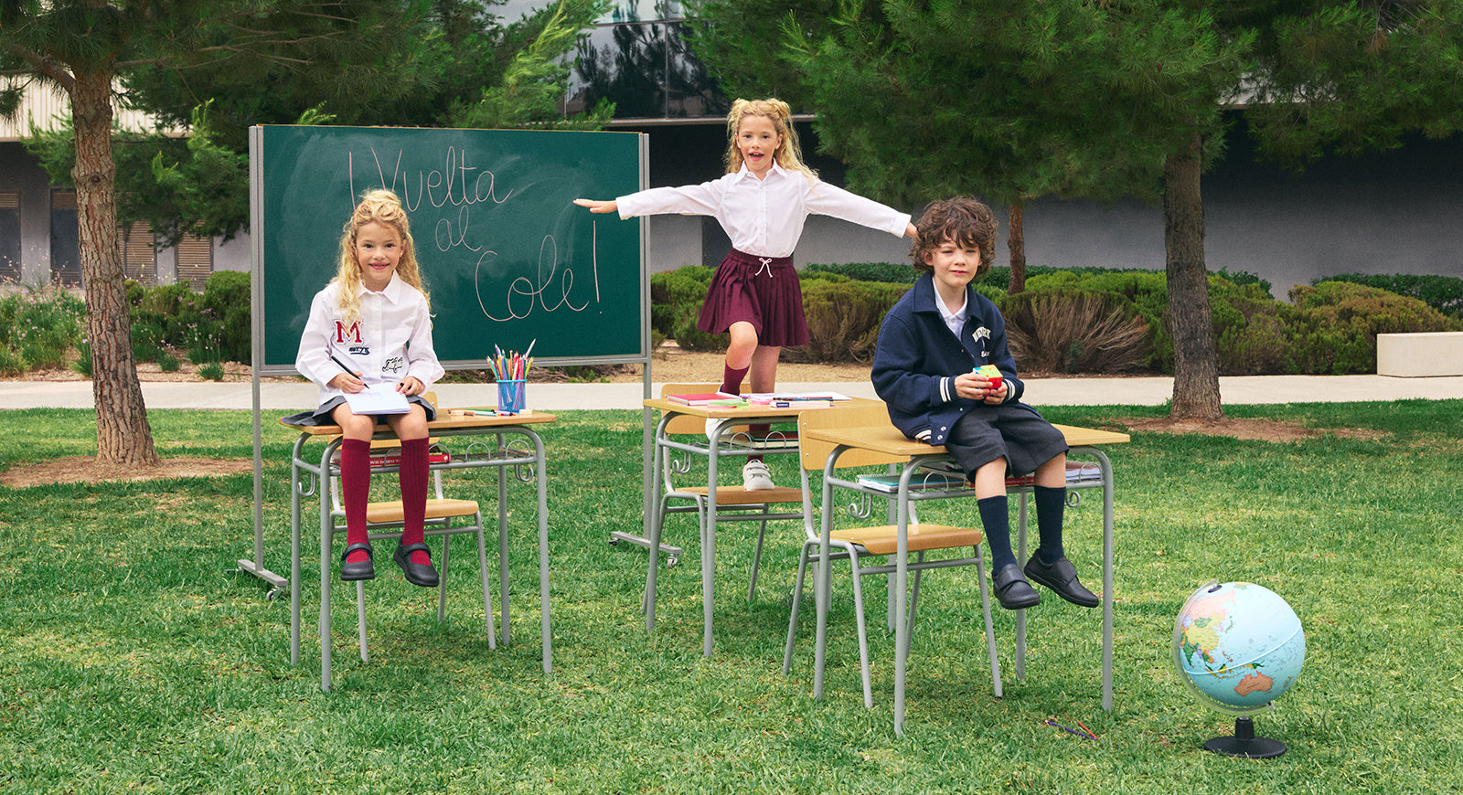 Tres niños con uniforme escolar en un aula improvisada al aire libre, frente a un pizarrón que dice "¡Vuelta al cole!", con escritorios, útiles y un globo terráqueo.