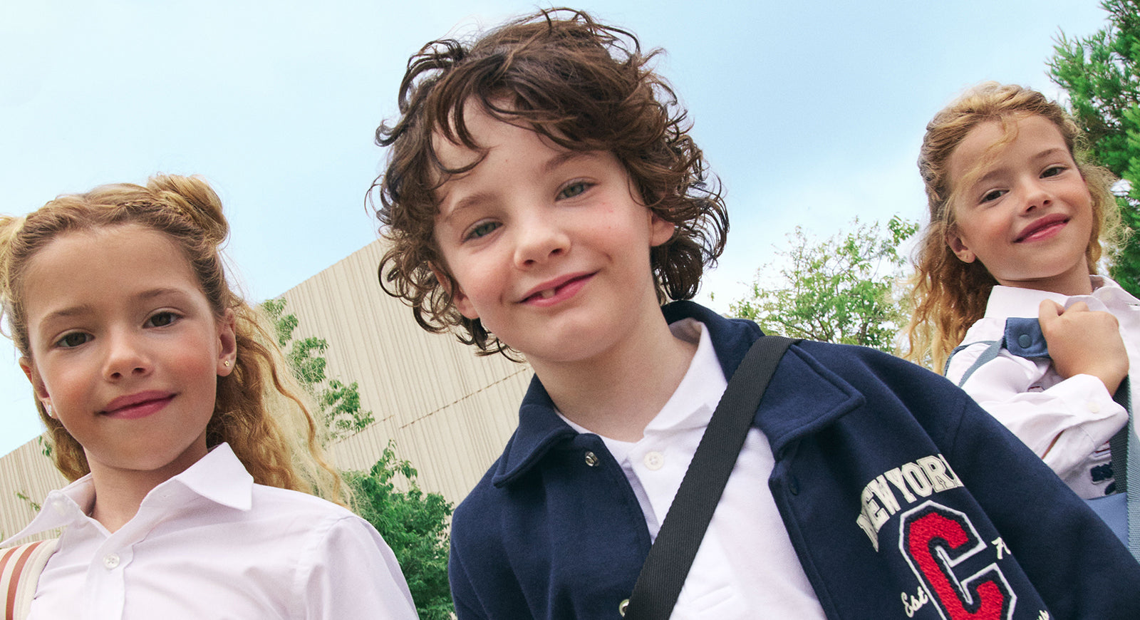 Tres niños sonrientes con uniforme escolar posando al aire libre con mochilas, frente a un edificio moderno y árboles verdes.