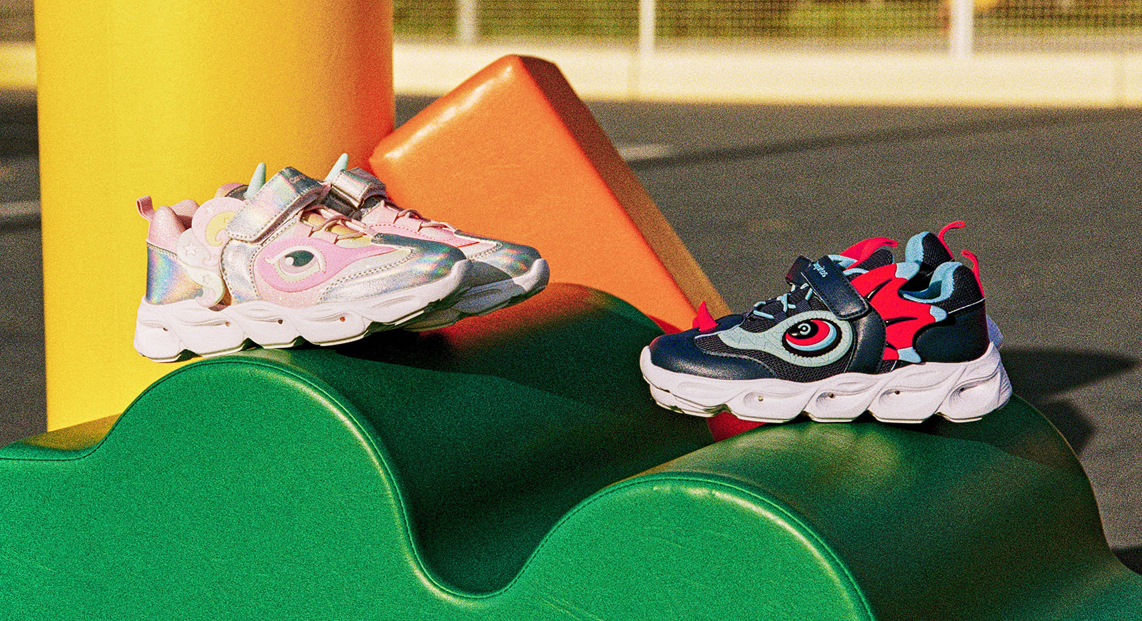 Zapatillas infantiles coloridas con diseños de criaturas fantásticas, una en tonos rosa y otra en azul marino, sobre un juego de plástico en un parque.