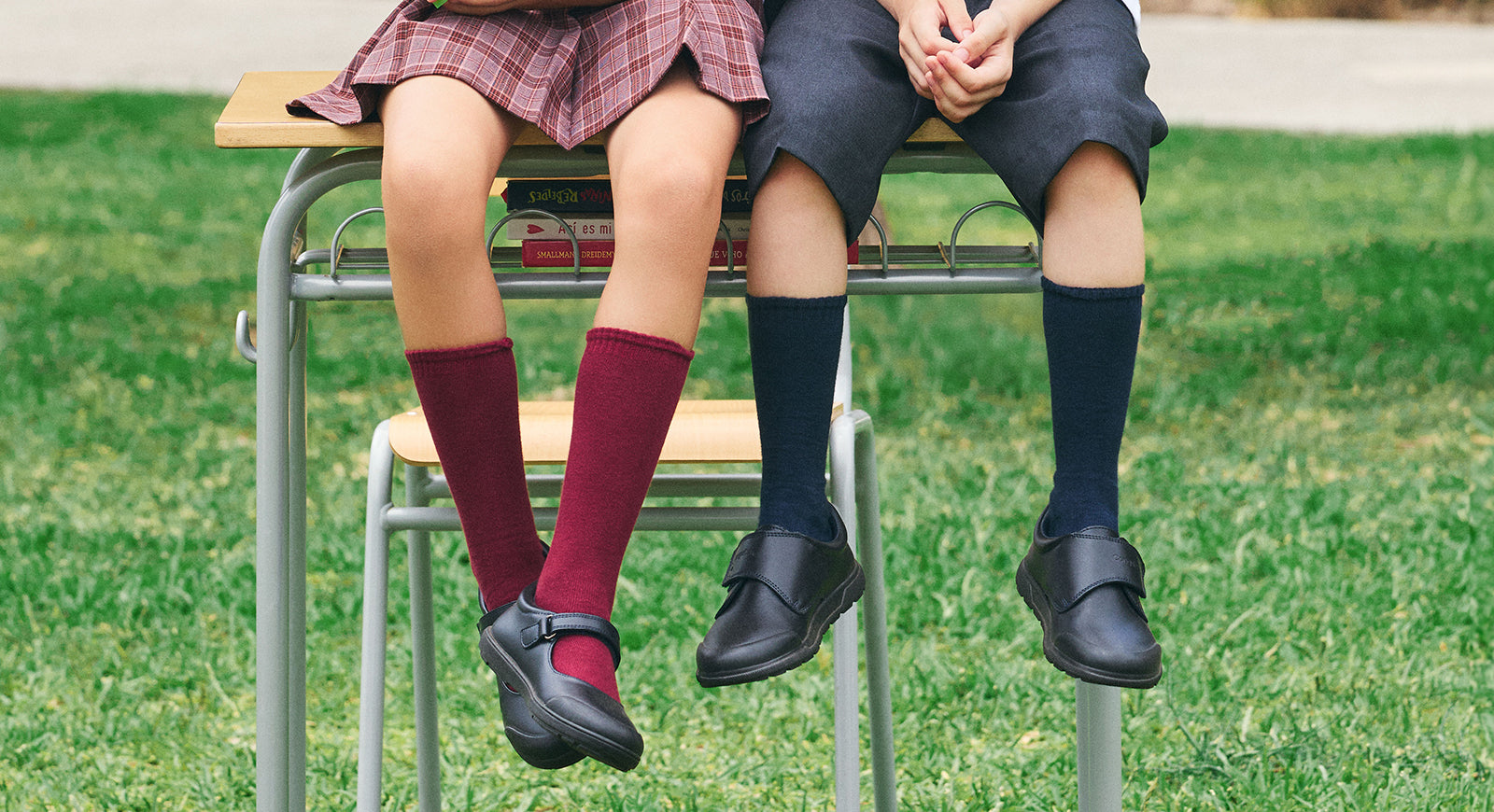 Niña y niño con uniforme escolar sentados sobre un escritorio, mostrando sus zapatos colegiales negros y calcetas largas burdeos y azul marino.